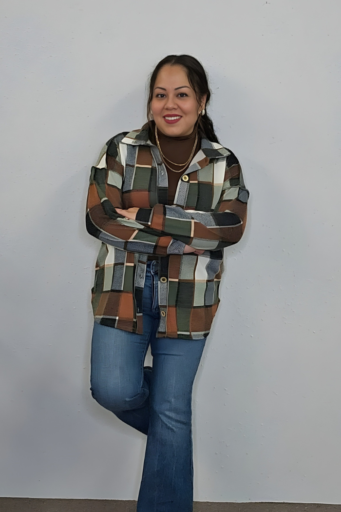 A woman with long dark hair smiles and poses with arms crossed, wearing the Plaid Print Shirt Jacket as a lightweight layer over a brown turtleneck, styled with blue jeans and hoop earrings against a plain light-colored wall.