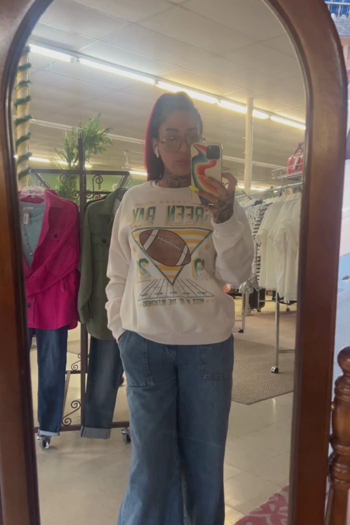 A person with red hair and visible tattoos is taking a mirror selfie video, wearing a cream-colored sweatshirt with a large graphic featuring a football and the text "GAME DAY" (reversed in the mirror), blue jeans, and holding a phone with a colorful case. The background shows clothing racks and other items in what appears to be a retail store or boutique.