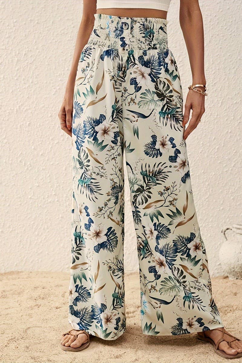 A person stands on sand in Tropical Wide Leg Pants featuring a tropical floral print and high-rise elastic waistband, paired with a matching crop top, tan sandals, and gold bracelets against a textured beige background.