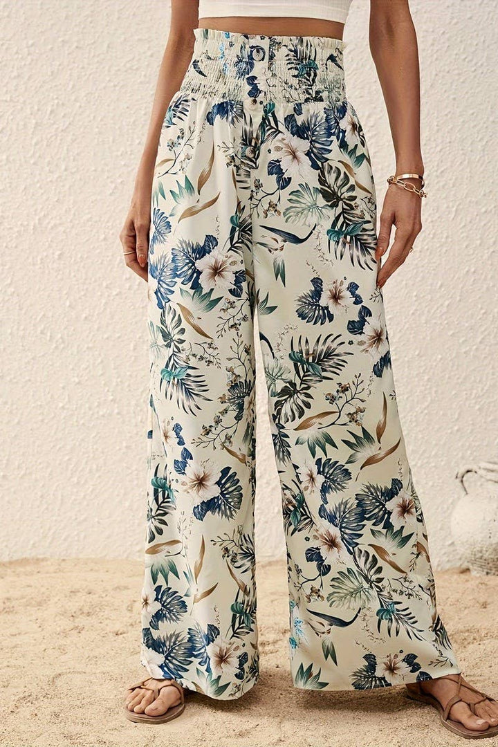 A person stands on sand in Tropical Wide Leg Pants featuring a tropical floral print and high-rise elastic waistband, paired with a matching crop top, tan sandals, and gold bracelets against a textured beige background.
