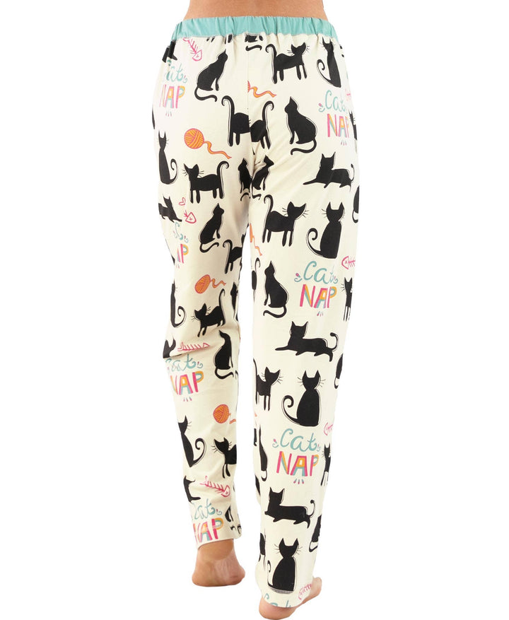 A person wears Dog Mom, Cat Nap & More Pajama Pants—cream-colored cotton pants with black cat silhouettes, yarn balls, and "Cats Nap" in colorful letters—shown from the back with bare feet.