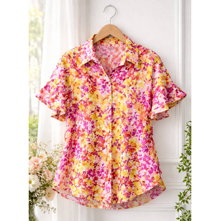 The Plus Floral Button Blouse, a vibrant pink, yellow, and orange floral print short-sleeve button-up for plus sizes, hangs on a wooden hanger against a white wall with flowers and greenery nearby.