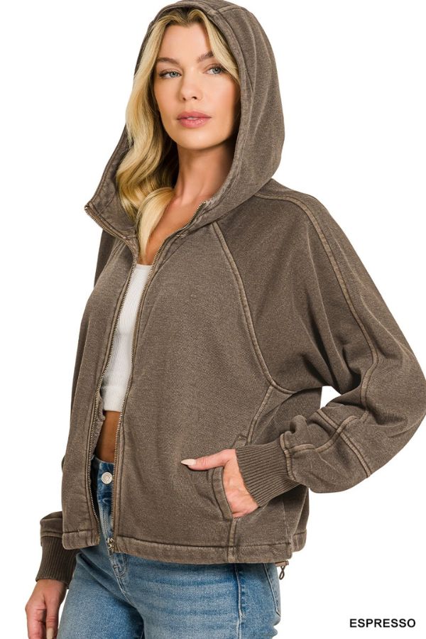 Woman wears the Pigment Dye Zip Up Hoodie in Espresso, featuring a brown zip-up fleece with adjustable hem, paired with a white crop top and blue jeans. She stands with one hand in her pocket, looking at the camera.