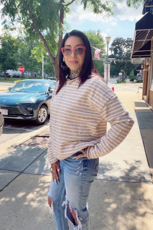 A woman stands on a sunny sidewalk wearing pink sunglasses, ripped jeans, and the Striped Button Front Sweater in beige and white, with parked cars and trees visible in the background.