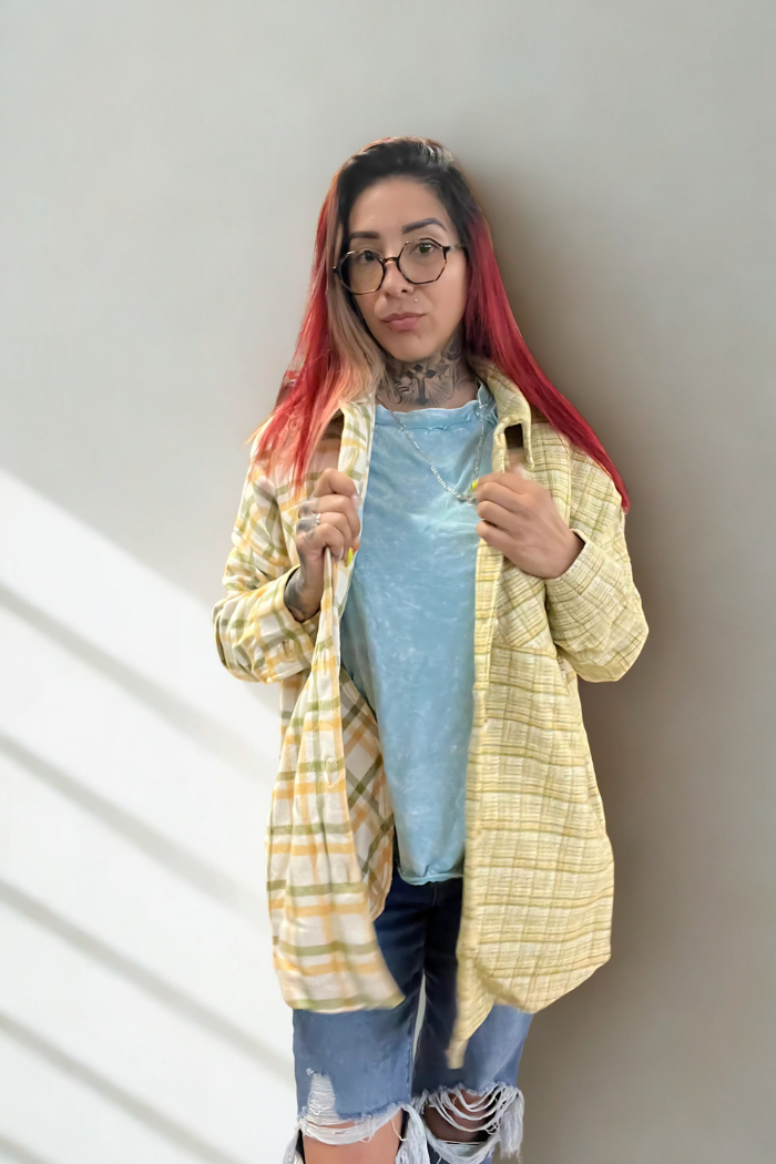 A person with red-tipped hair and glasses stands facing forward, wearing a light blue t-shirt under an open yellow, white and green plaid shacket, with visible tattoos on their neck and chest, and ripped blue jeans.