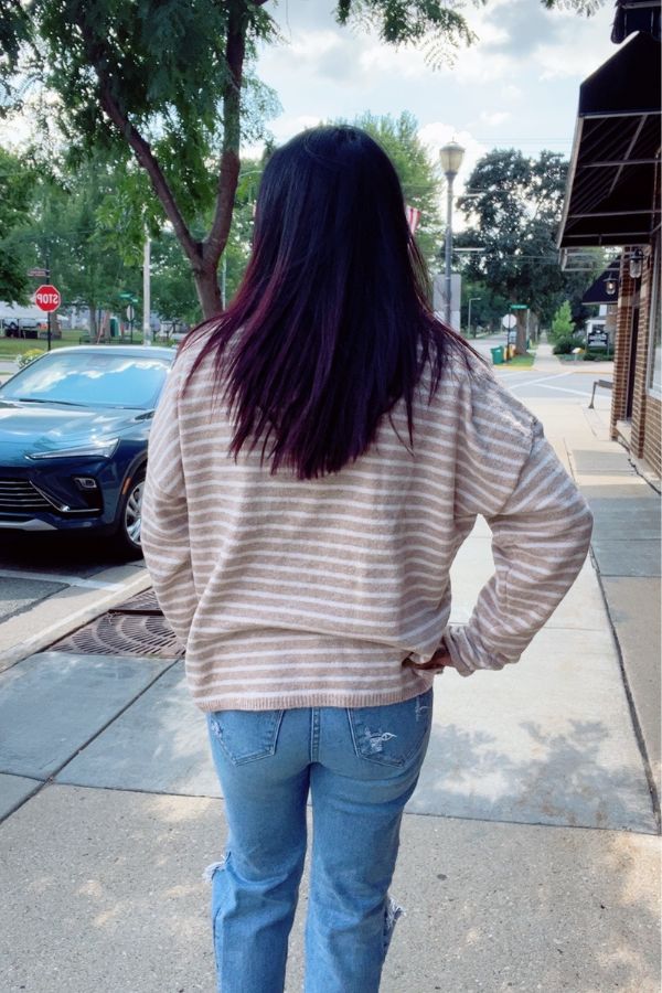 A person with long dark hair, wearing a Striped Button Front Sweater and blue jeans, stands on a sidewalk with their back to the camera. Trees, parked cars, and buildings are visible in the background.