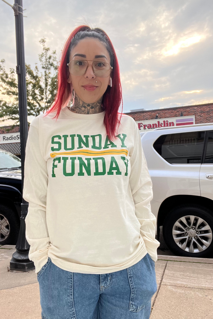 A person with red hair and glasses wearing a long-sleeved cream-colored shirt with "SUNDAY FUNDAY" in green and yellow text, and blue jeans, standing outdoors in front of buildings. 