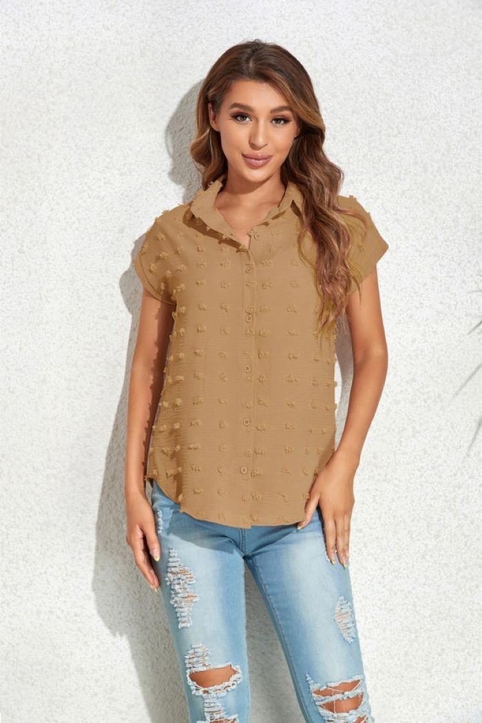 A woman with long, wavy brown hair stands by a light textured wall, wearing a short-sleeve Textured Button Blouse and light blue ripped jeans. She looks relaxed with one hand in her pocket.