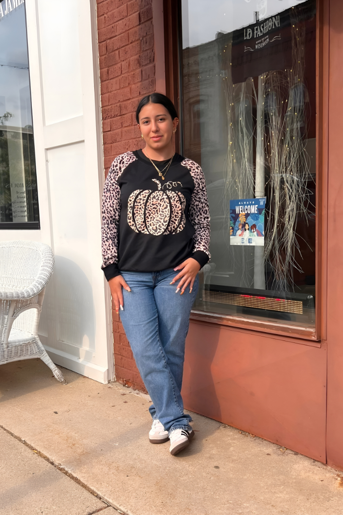 A young woman with dark hair stands on a sidewalk in front of a brick building with a large storefront window. She is wearing a black long-sleeved shirt with a leopard print pumpkin design on the front and leopard print sleeves, blue jeans, and white sneakers with black stripes.