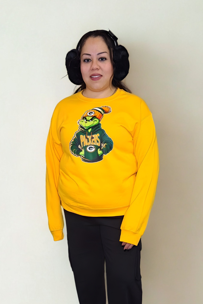 A woman with dark hair in side buns wears the Green Bay Packers Grinch Sweatshirt and black pants, posing against a plain light background for a playful holiday crewneck look.