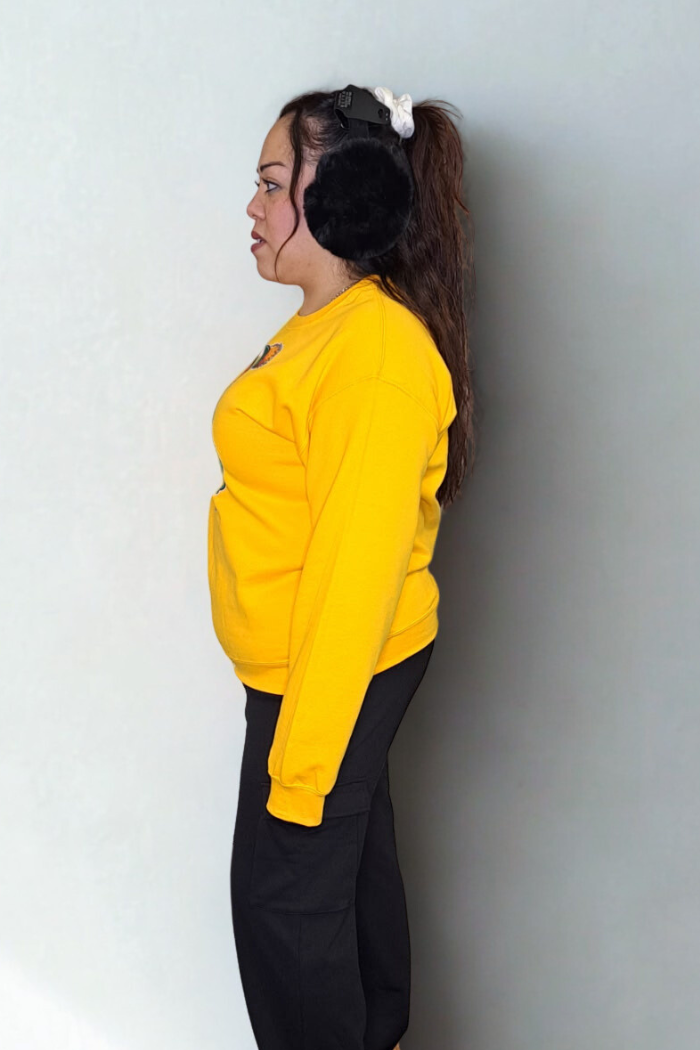 A woman stands in profile against a light background, wearing a Green Bay Packers Grinch Sweatshirt, black pants, fluffy black earmuffs, and a white scrunchie in her ponytail.