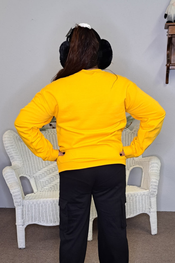 A person stands with hands on hips, back to the camera, wearing a bright yellow Green Bay Packers Grinch Sweatshirt, black pants, and black earmuffs. A white wicker bench and a light gray wall are visible in the background.