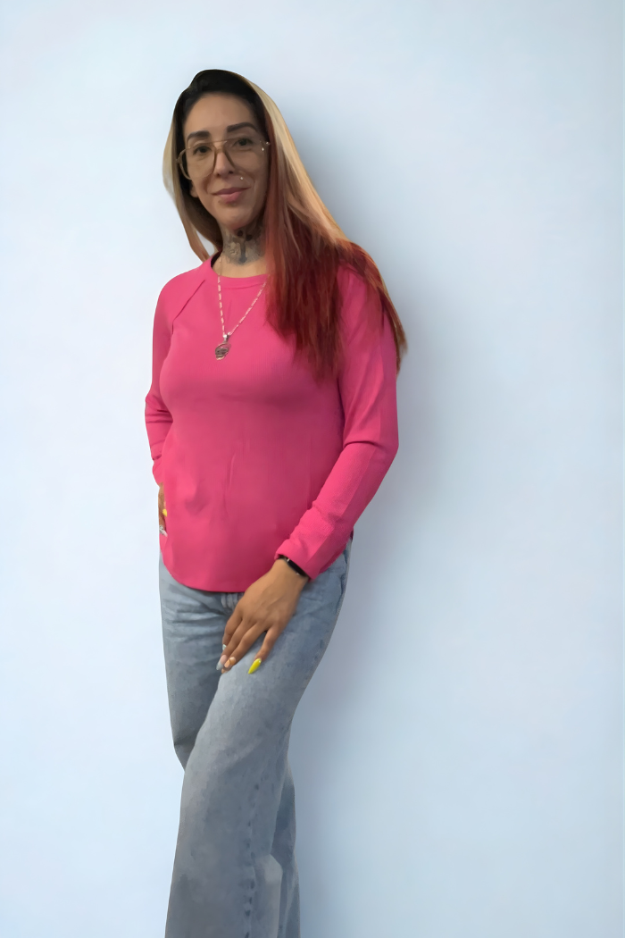 A person with long brown hair and glasses smiles with one hand in their pocket, wearing the Waffle Knit Long Sleeve Top in pink, light blue jeans, and a necklace, standing against a plain light background.