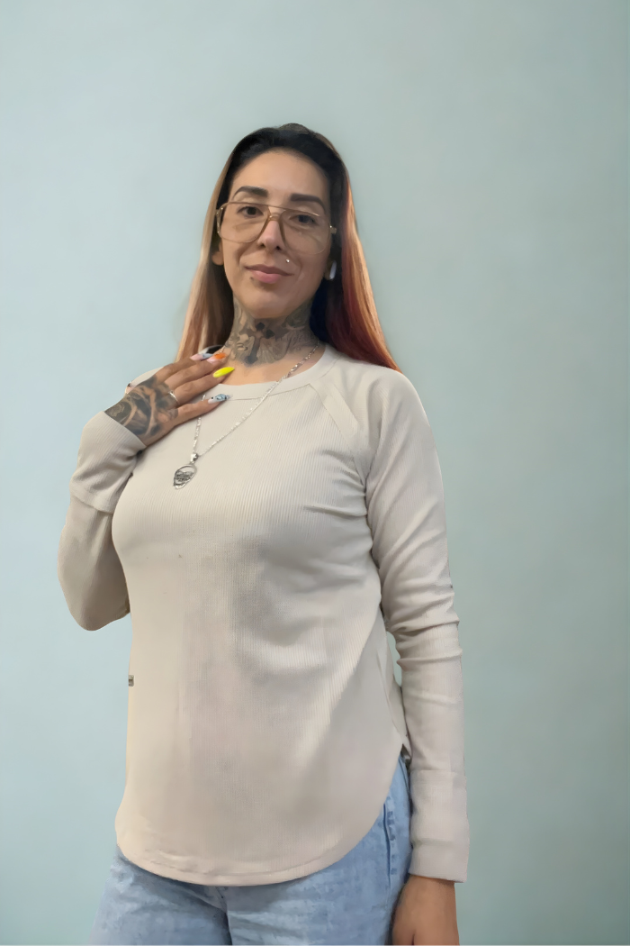 A woman with long hair, glasses, tattoos, and painted nails wears a Waffle Knit Long Sleeve Top with blue jeans. She stands against a light blue background, smiling slightly and touching her necklace.