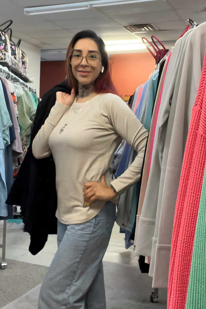 A woman with glasses and long hair poses confidently in a clothing store, wearing a Waffle Knit Long Sleeve Top and light blue jeans, with a dark jacket over her shoulder and colorful clothes on racks around her.