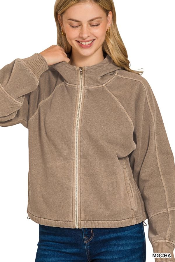 Woman wearing the Pigment Dye Zip Up Hoodie in mocha with a relaxed fit and adjustable hem, smiling with one hand near her collar; she has long blonde hair and blue jeans.