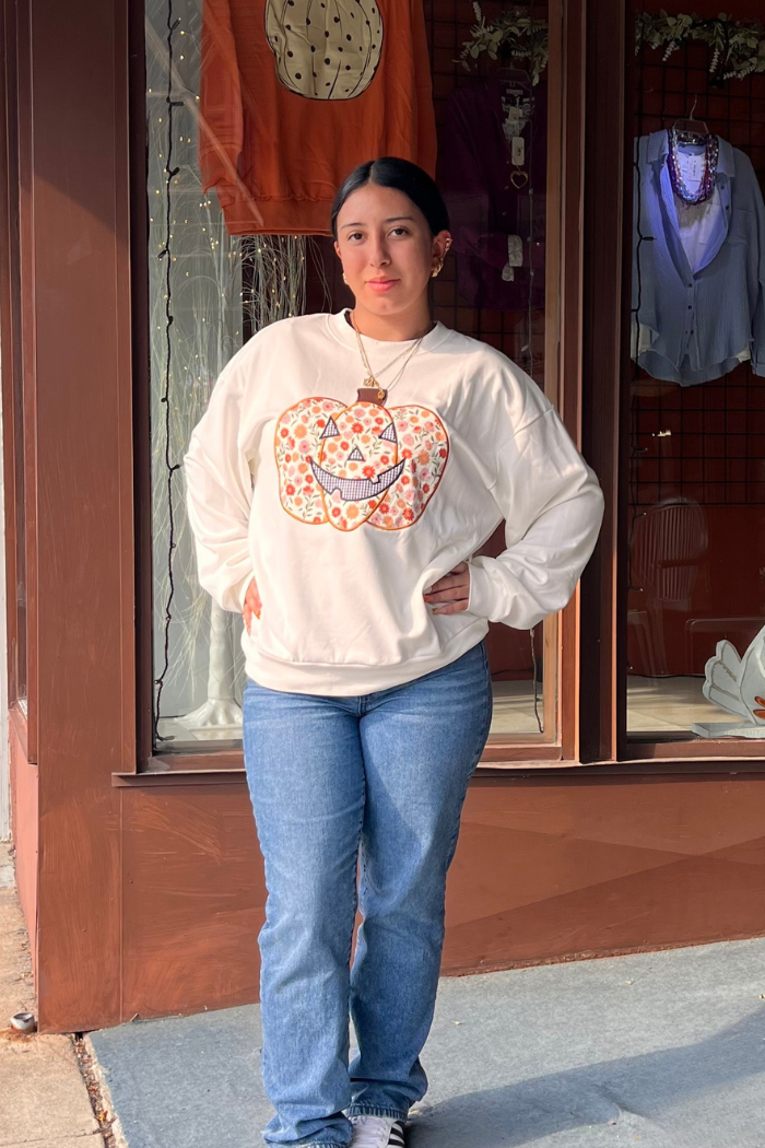 A young woman stands in front of a window, wearing a white sweatshirt adorned with a floral print jack-o'-lantern design and blue jeans. She has dark hair pulled back, a necklace, and is posing with her hands on her hips, looking directly at the viewer. 