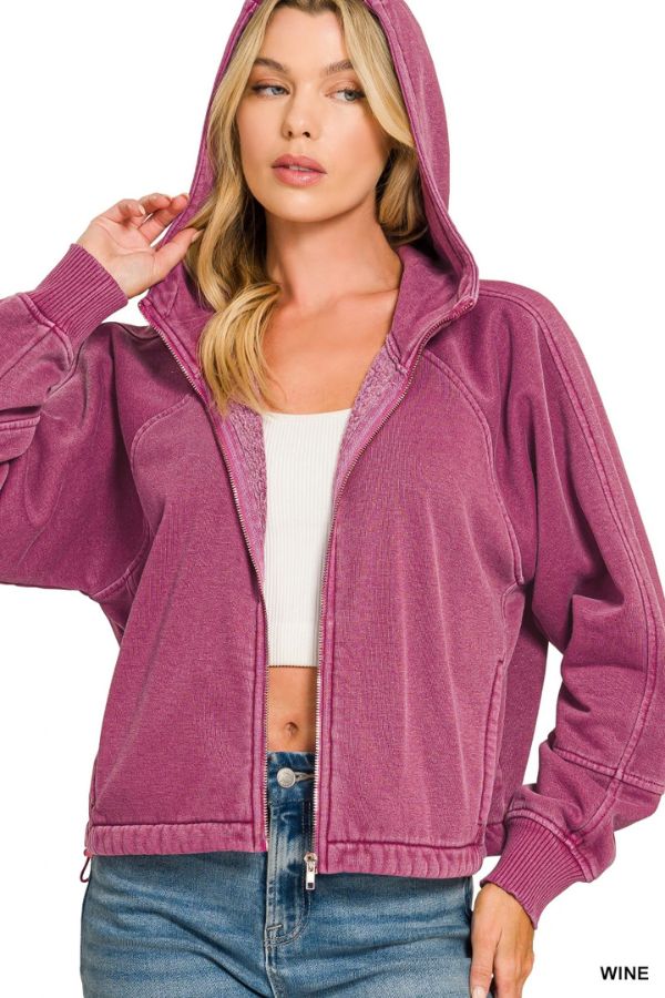 Woman in a wine-colored Pigment Dye Zip Up Hoodie with hood and adjustable hem over a white crop top and blue jeans, holding the edge of the hood with one hand.