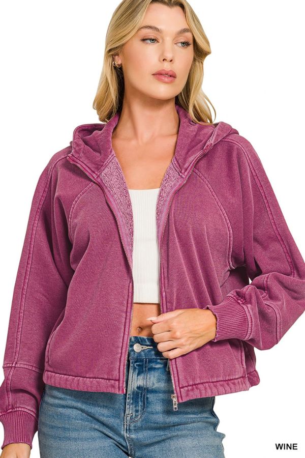 A woman in a wine-colored Pigment Dye Zip Up Hoodie with an adjustable hem over a white crop top and blue jeans stands looking slightly to the side.