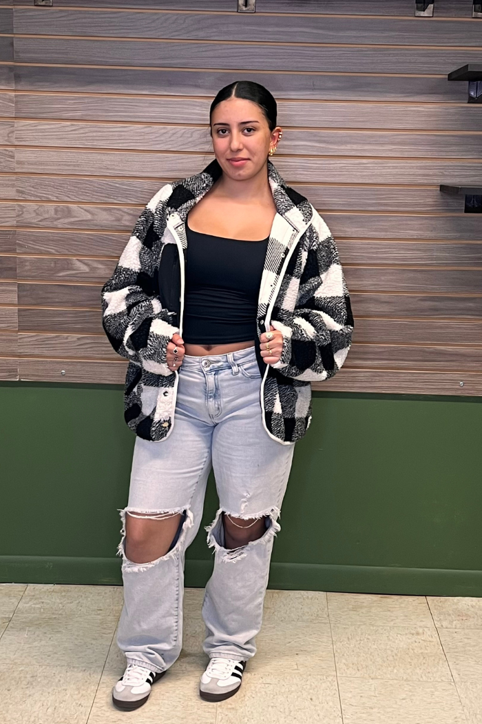 A young woman stands facing forward, wearing a black and white checkered plaid shacket or jacket over a black crop top. She is also wearing light-wash, ripped wide-leg jeans and white sneakers with black stripes. Her dark hair is pulled back, and she has visible piercings in her ears and nose.