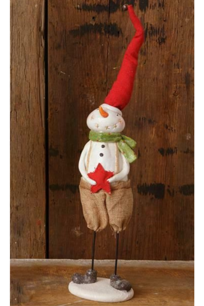 A primitive-style Christmas snowman figurine stands on a white base against a rustic wooden background. The snowman has a white, textured body, a long, pointed red hat with a small bell at the tip, a green scarf, and burlap-like short pants. It has thin, dark wire legs ending in brown boots and holds a red star in its hands. 