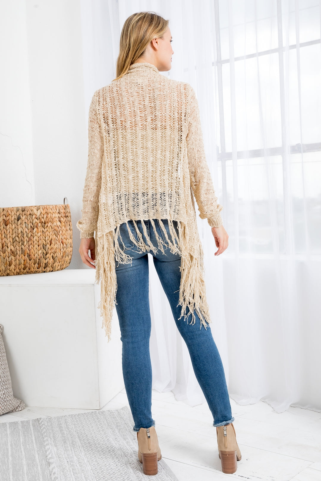 Woman wearing a beige cardigan with fringe, blue jeans, and brown boots in a bright room.