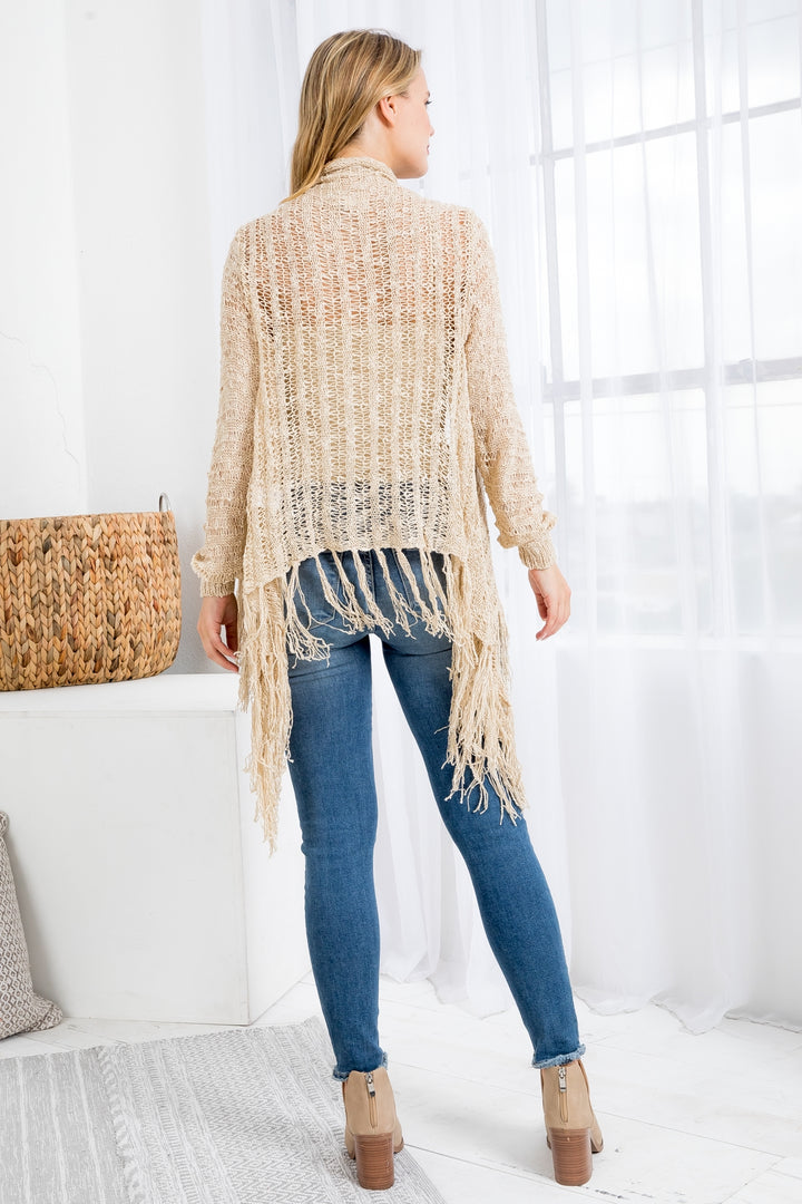 Woman wearing a beige cardigan with fringe, blue jeans, and brown boots in a bright room.