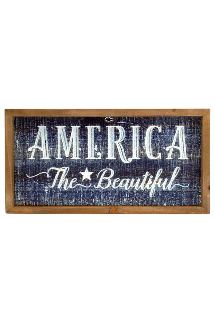 A rectangular wooden sign with a distressed dark blue background and white text. The text reads "AMERICA" in a large, blocky font, followed by "The Beautiful" in a smaller, script font with a star in between the and beautiful.