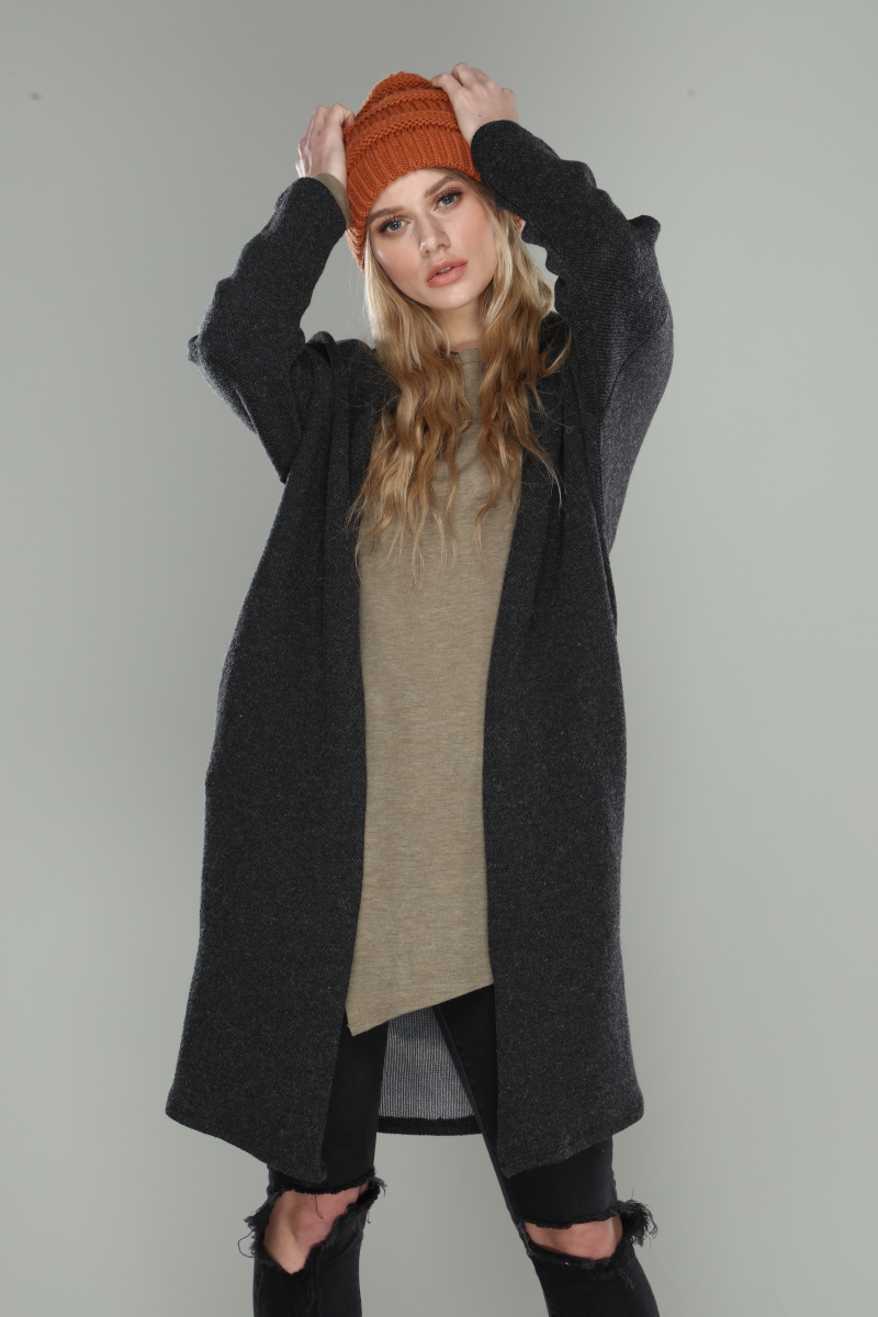 Woman wearing a long dark coat, beige sweater, and orange beanie against a plain background