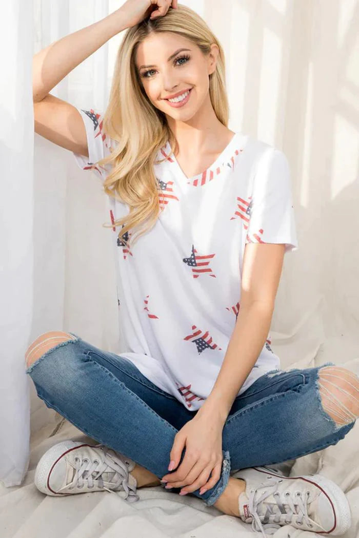 A smiling young woman with long blonde hair sits cross-legged on a light-colored surface, leaning against a white curtain. She is wearing a white V-neck t-shirt with red and blue sequin stars and stripes, and distressed blue jeans with ripped knees. She is also wearing white sneakers. 