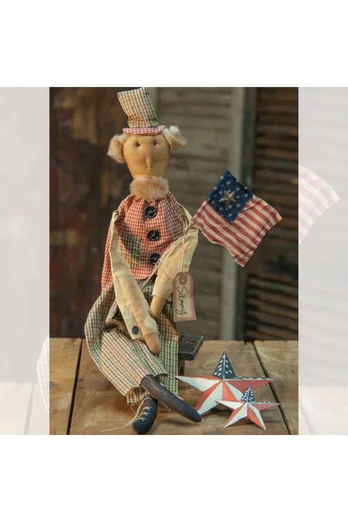A primitive-style Uncle Sam doll, known as the "Sammy Doll," is seated on a wooden surface. The doll wears a red gingham vest with black buttons, green plaid pants, and a matching plaid top hat. It holds a homespun American flag with a tin star attached. Two primitive, red, white, and blue star-shaped decorations are placed in front of the doll. 