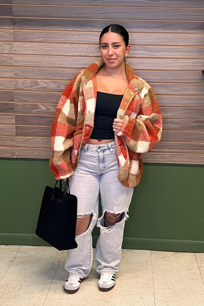 A young woman stands facing forward, wearing a patterned orange, brown, and off-white plaid sherpa jacket over a black crop top. She pairs this with light-wash, distressed jeans featuring large rips at the knees, and white sneakers with black stripes. She holds a black tote bag in her left hand.