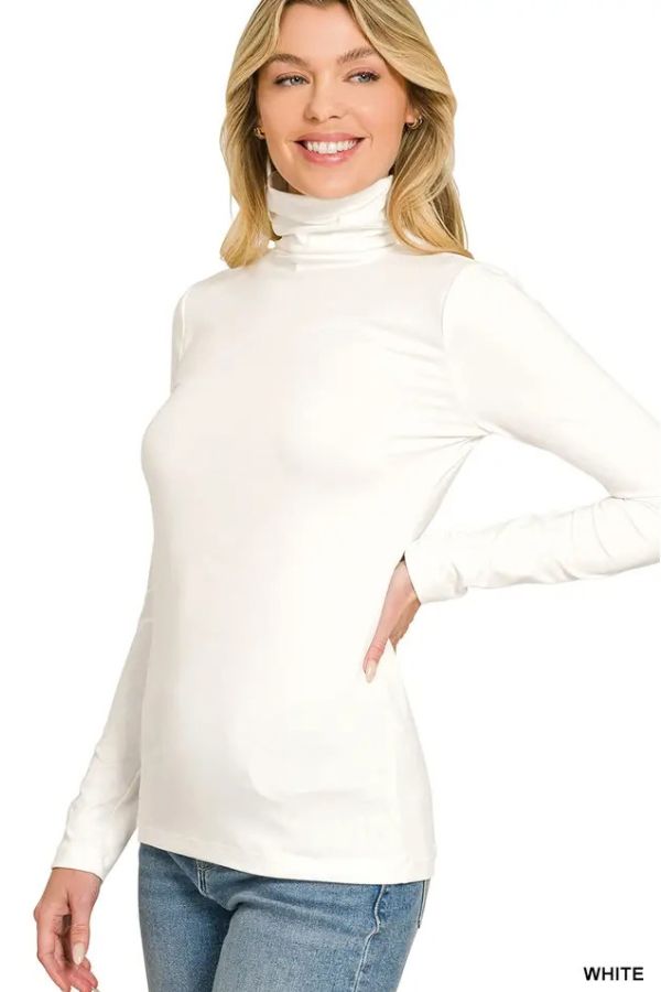 A woman smiles against a plain white background, standing with one hand on her hip while wearing the Premium Microfiber Mock Neck Top in white paired with blue jeans.
