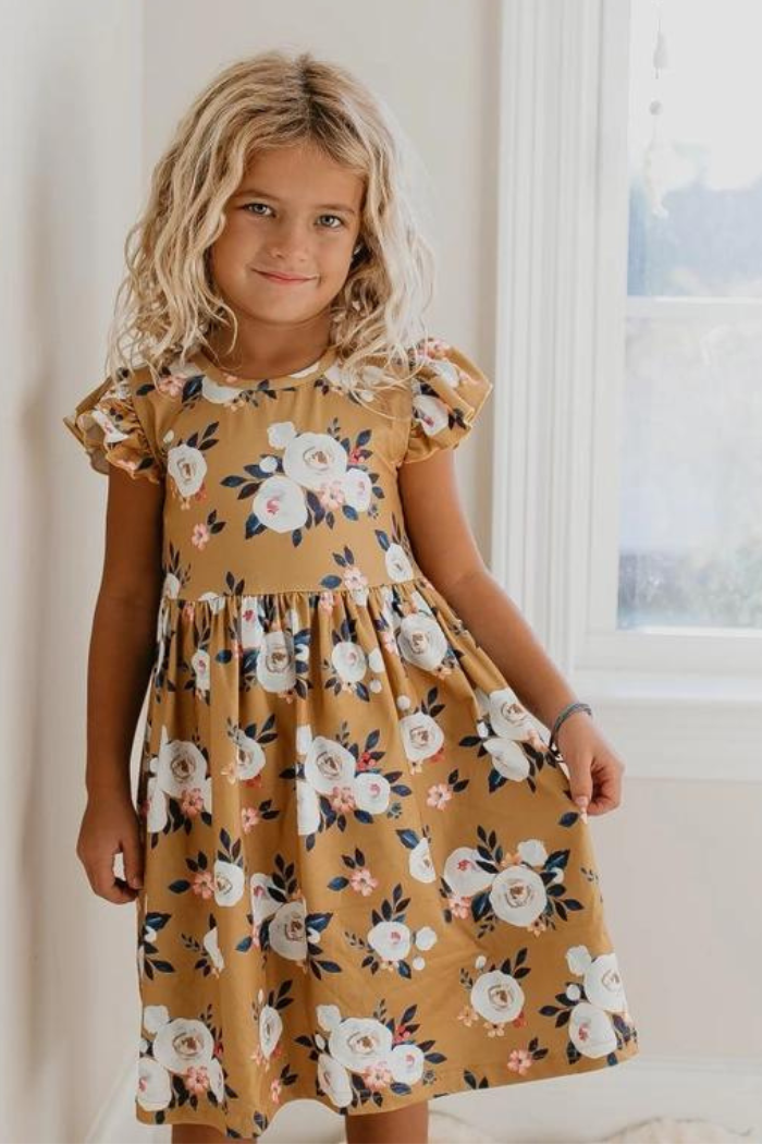 The dress is mustard yellow with a floral pattern of white and pink roses with green leaves. It has short, ruffled sleeves and a gathered waist.