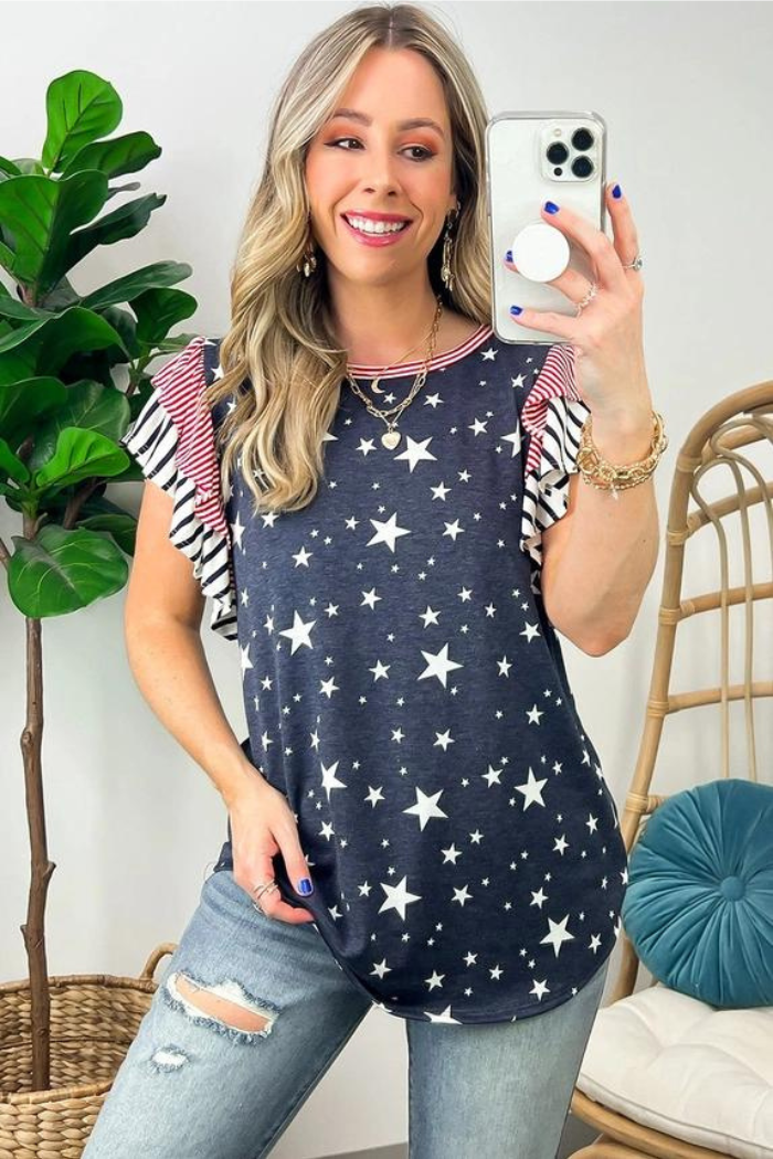 A woman wearing a patriotic-themed shirt with white stars and red and white striped ruffled sleeves. She's also wearing jeans and has multiple pieces of jewelry. The shirt is likely intended for Independence Day celebrations.