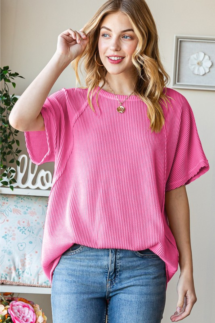 A smiling woman with wavy blonde hair wears the Solid Urban Ribbed Top with dolman short sleeves and blue jeans. She stands indoors by a floral pillow and plant, touching her hair.