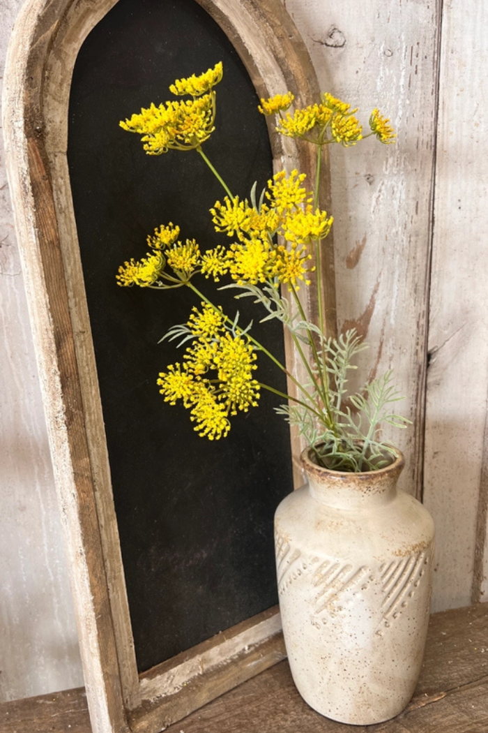 The image shows a small, textured vase containing several stems of bright yellow flowers artificial Queen Anne's Lace, set against a rustic backdrop. 