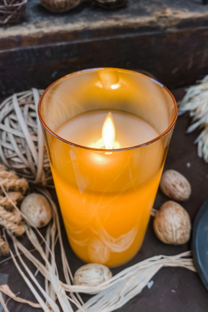The image shows an amber glass cylinder moving flame candle, surrounded by decorative elements such as woven twine, small nuts, and straw-like strands.