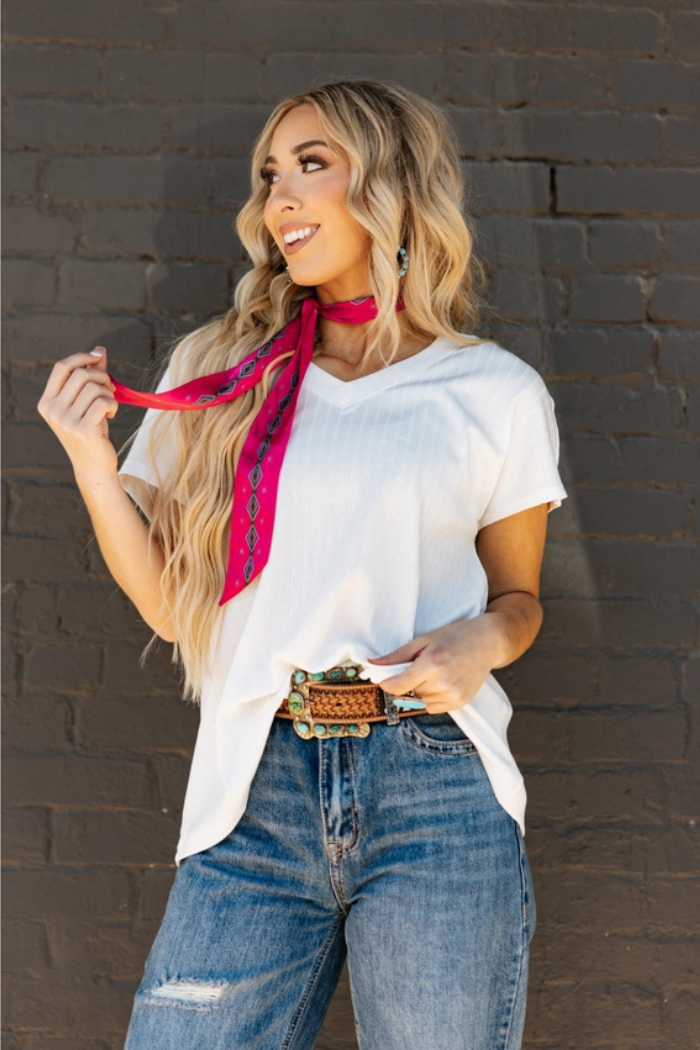 Woman wearing a plus size ribbed v neck top in ivory with relaxed fit and cozy ribbed texture, blue jeans, and a pink scarf against a brick wall.