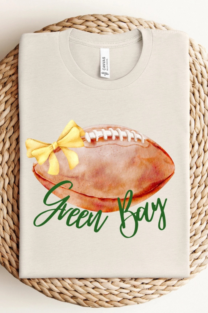 T-shirt with a design featuring a watercolor-style football, a yellow ribbon, and the text "Green Bay" in cursive. 