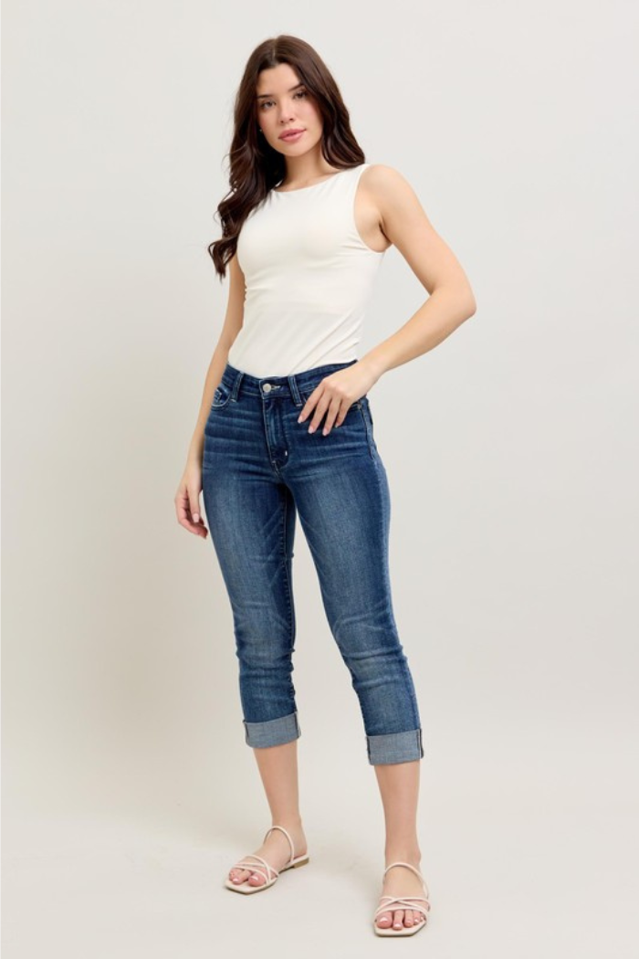 Woman modeling a pair of Judy Blue jeans, specifically the "Cuffed Capri" style. She is wearing a white tank top and flat sandals, posing against a plain background. The jeans are a medium wash with cuffed hems, ending just below the knee. Judy Blue is known for its comfortable and stretchy denim, often praised for its fit and softness.