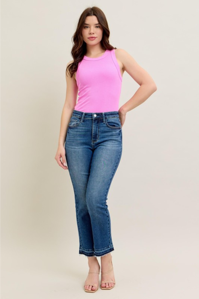 The image shows a woman modeling a pair of Judy Blue jeans, specifically the "Release Hem Crop Kick Flare" style. She is wearing a bright pink tank top and open toe heeled sandals. The jeans are a medium-wash denim with a cropped flare leg and a released hem, meaning the raw edge of the hem is visible. They appear to be a high-waisted style, fitting snugly at the waist and hips. The model is standing with one hand on her hip, facing forward, against a plain background.