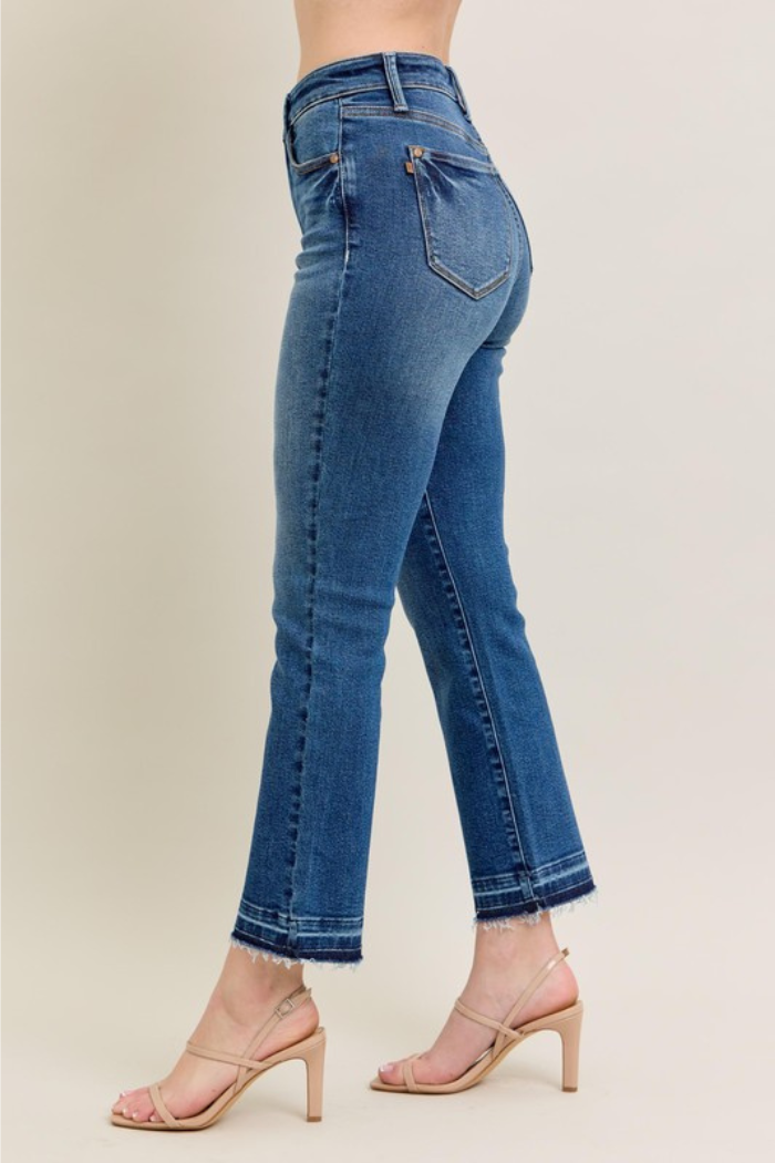 The image shows a pair of Judy Blue High Waist Release Hem Kick Flare Jeans in a medium wash denim. The jeans feature a high waist, a kick flare leg, and a released hem with frayed detailing. The model is also wearing nude-colored heeled sandals. Judy Blue is known for its comfortable and flattering denim, designed to hold its shape and fit a variety of body types.