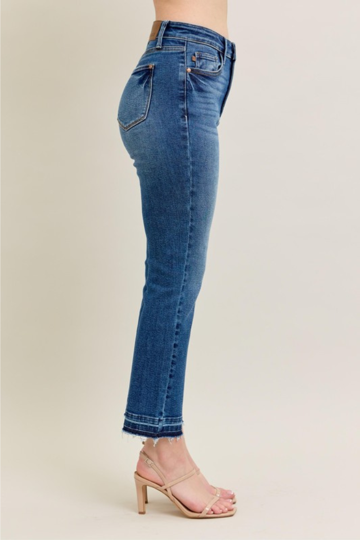 The image shows a person wearing Judy Blue High Waist Release Hem Kick Flare Jeans. These jeans are characterized by a high waist, a slim fit through the thighs, and a subtle flare starting below the ankle. The hem features a released, frayed edge, adding a touch of casual detail. The wash is a classic medium blue denim, and the jeans are paired with nude strappy heels.