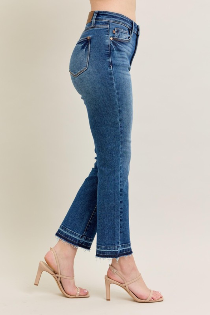 The image shows a pair of Judy Blue High Waist Release Hem Kick Flare Jeans in a medium wash denim, worn by a model. These jeans feature a high waist, a kick flare leg opening, and a released hem, which gives them a slightly frayed and unfinished look. The model is also wearing nude-colored heeled sandals. Judy Blue is known for its comfortable and stretchy denim. The kick flare style is a modern take on the classic flare jean, offering a subtle flare from the knee down. 