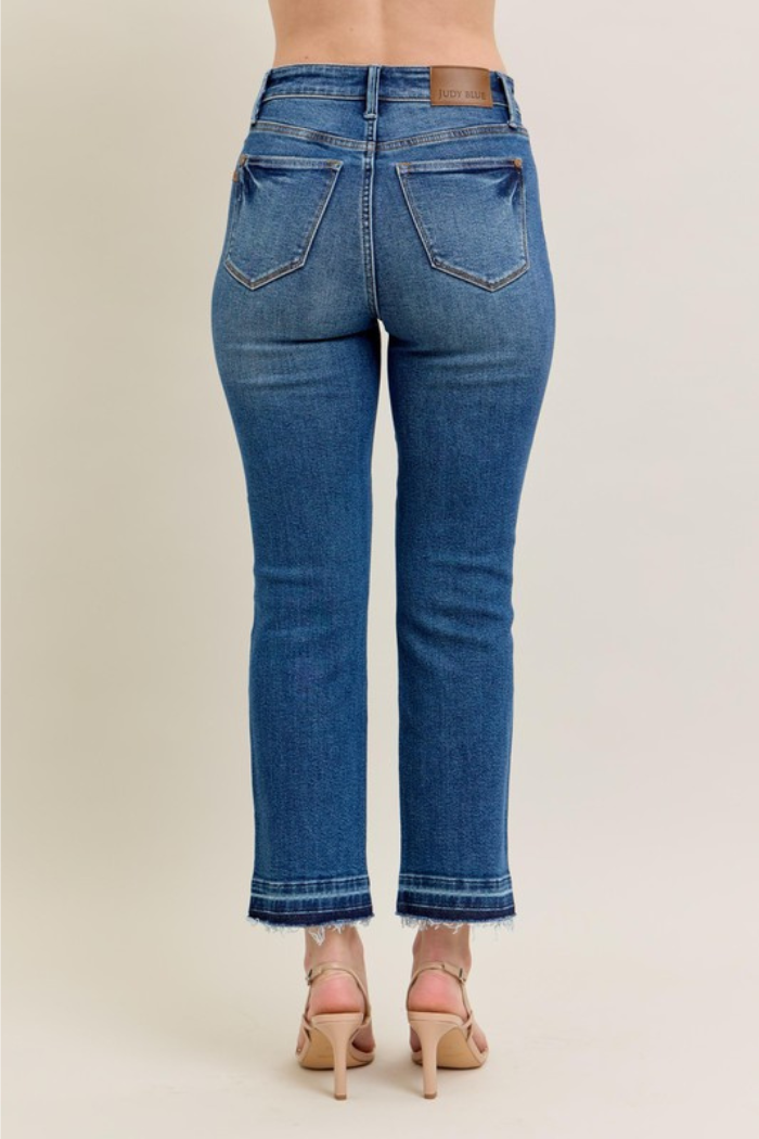 The image shows the back view of a person wearing Judy Blue high-waisted jeans and nude heels. The jeans are a medium wash with a frayed hem and the brand's leather patch visible on the waistband.