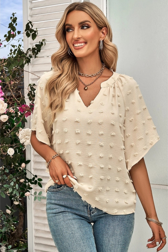 Woman wearing a cream-colored blouse with textured details and a V-neck. She is also wearing jeans, layered necklaces, and hoop earrings. The background appears to be a decorative wall or door with floral elements. The blouse is a light, neutral color with a relaxed fit, suitable for various occasions.