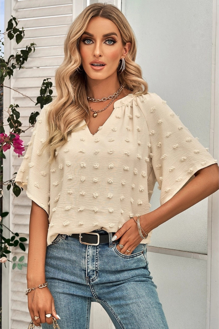Woman wearing a cream-colored blouse with small, raised dots, paired with blue jeans and a black belt. She has long blonde hair and is accessorized with a necklace, earrings, and bracelets. The background appears to be a white shuttered wall with some greenery and pink flowers visible on the left side. The style of the blouse and the overall composition suggests a casual, fashionable look.