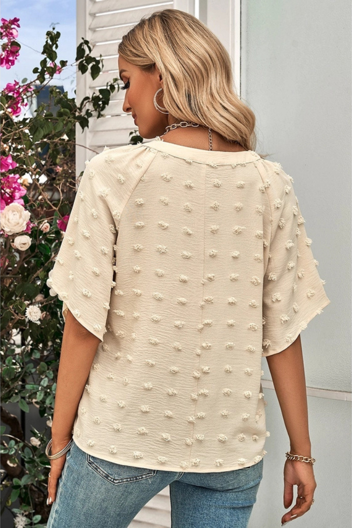 Woman is wearing a Swiss Dot Sleeve Blouse. The blouse is from the back it's light beige color and flared sleeves. It is covered in small, raised dots, giving it a textured look. The woman is also wearing jeans and some jewelry.