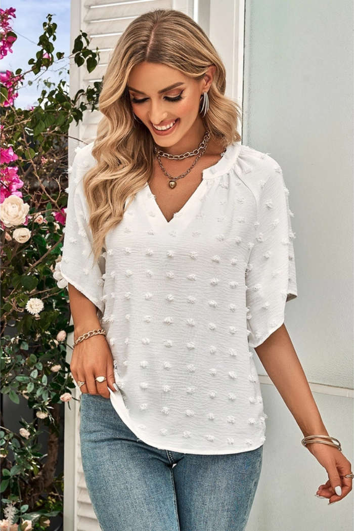 Woman wearing a white-colored blouse with small, raised dots, paired with blue jeans and a black belt. She has long blonde hair and is accessorized with a necklace, earrings, and bracelets. The background appears to be a white shuttered wall with some greenery and pink flowers visible on the left side.